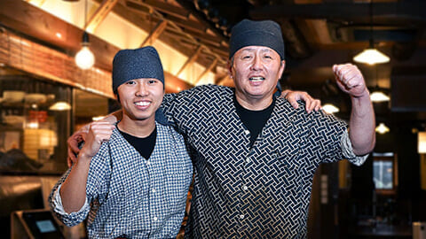 炭火焼き酒房すいしんの学生アルバイトと男性正社員が肩を組んで笑顔を向けている。